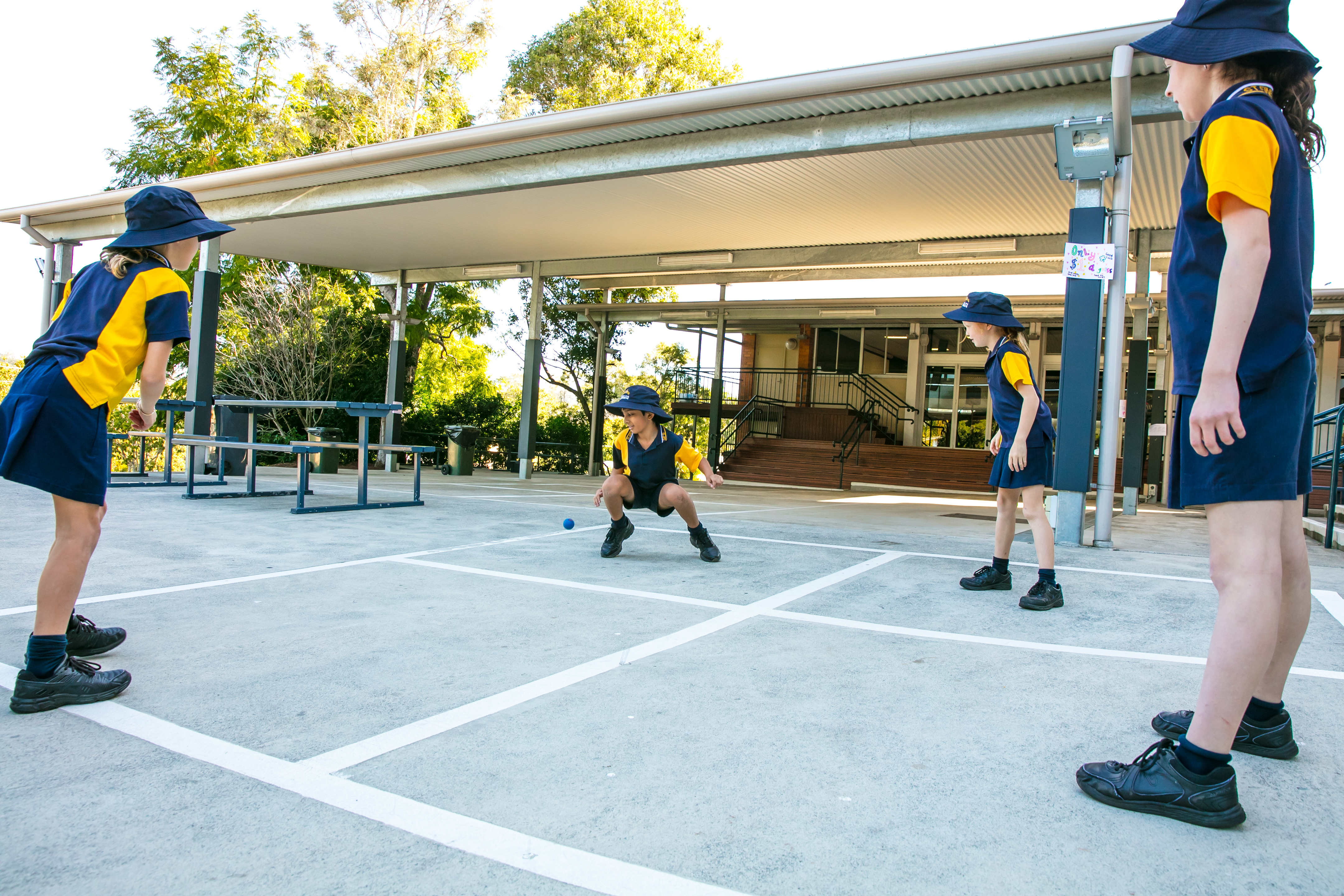 Students playing a playground ball game in a marked court under a covered area.