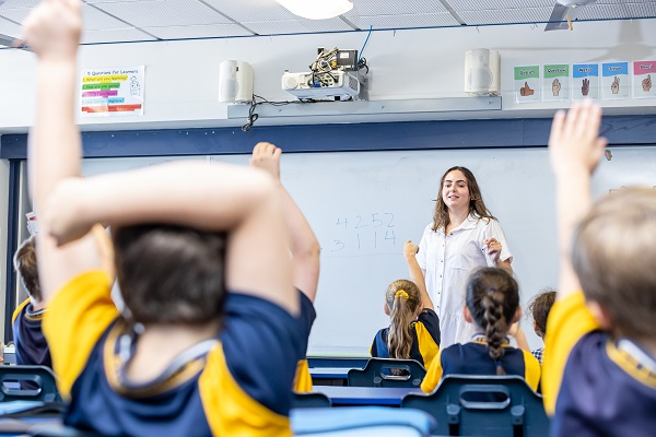 Teacher standing at the front of a classroom while students raise their hands during a lesson.