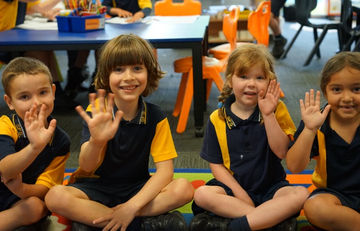 Young students in school uniform sitting on a classroom mat and raising their hands.