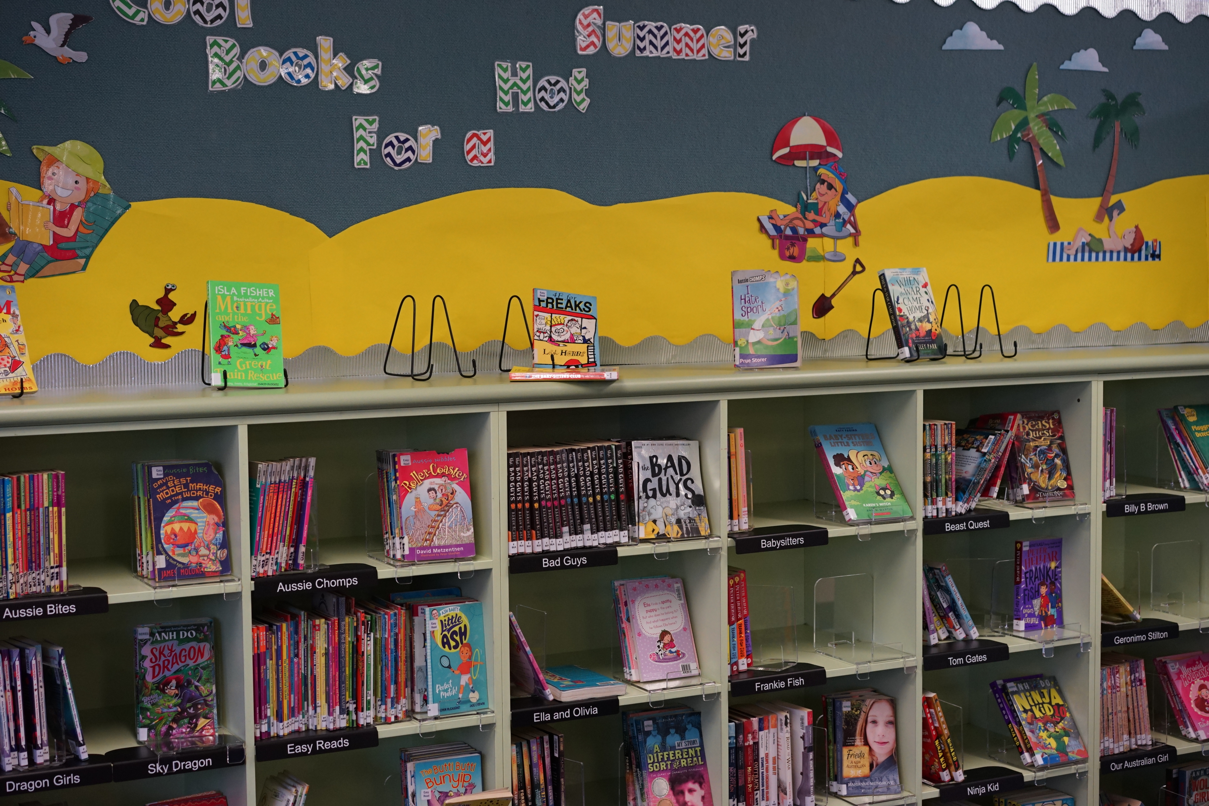 School library shelves displaying children’s books, with a summer‑themed book display above.