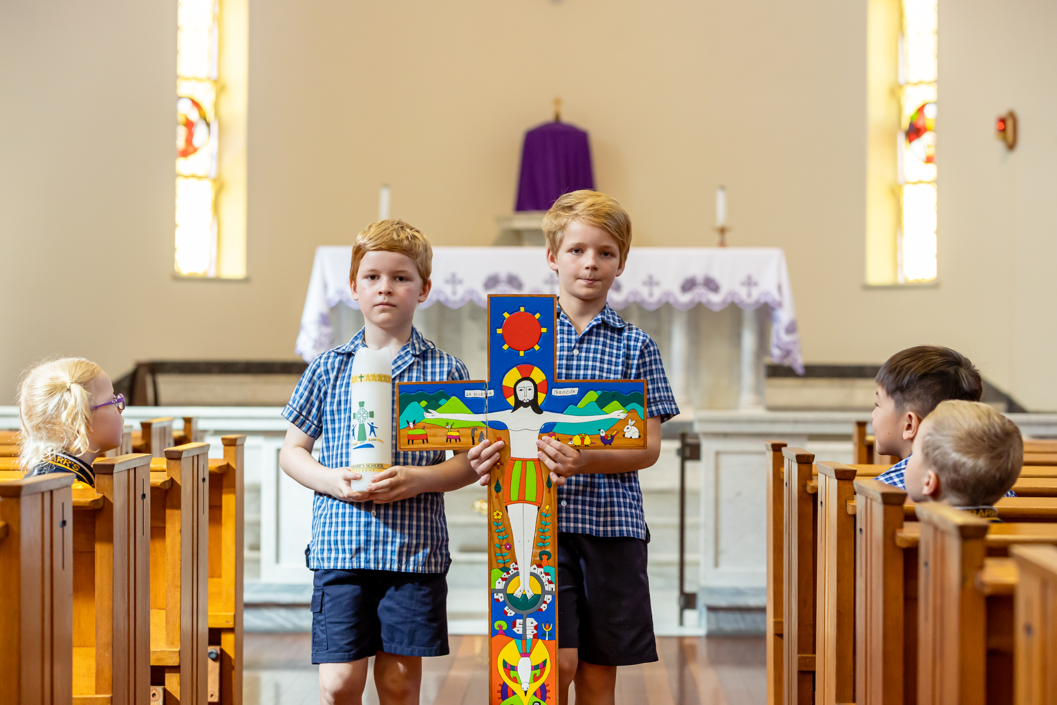 Two students in school uniform standing at the front of a church holding a cross and a candle.