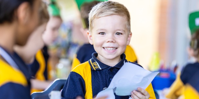 Student seated at a table holding paper during a classroom craft activity.