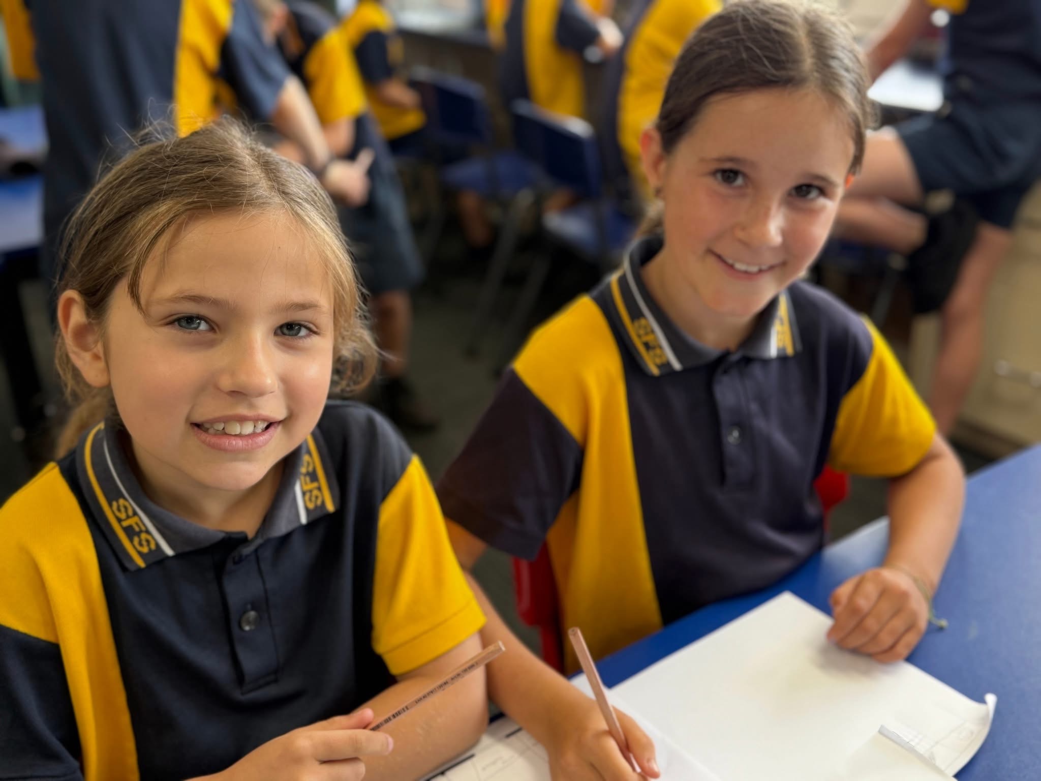Two students in school uniform seated at desks, writing during a classroom lesson.