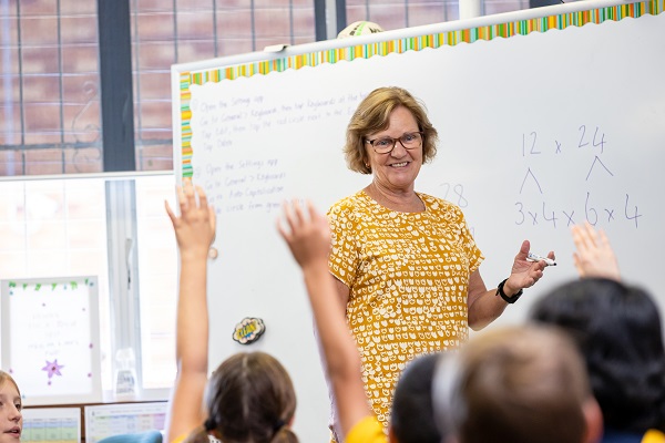 Teacher standing at a whiteboard while students raise their hands during a classroom lesson.