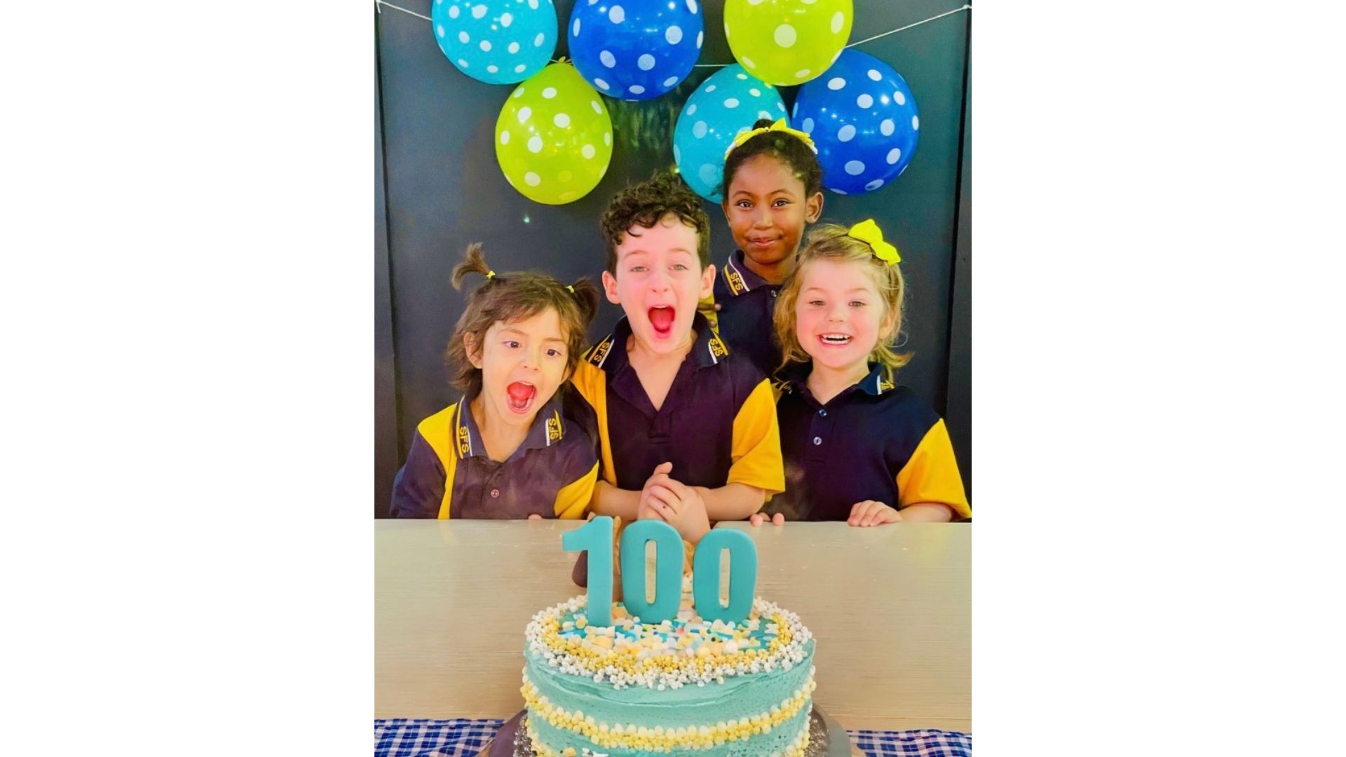 Students in school uniform standing behind a celebration cake topped with the number 100 and balloons.
