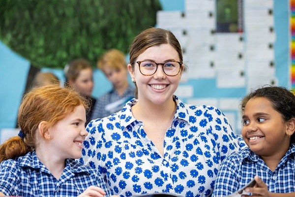 Adult seated with two students in school uniform during a classroom activity.