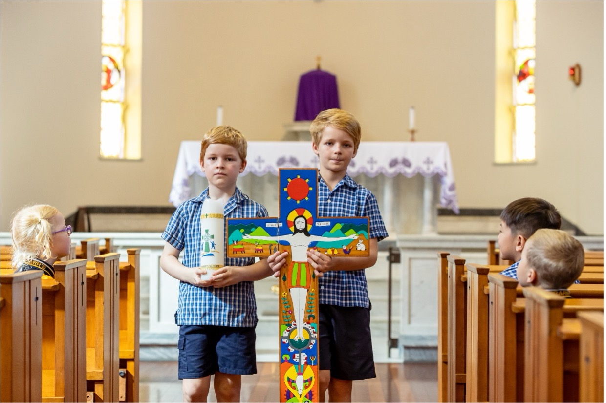 Two students in school uniform standing at the front of a church holding a decorated cross and a candle.